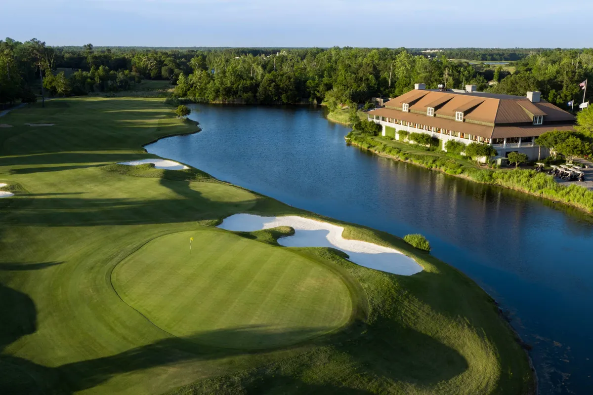 A scenic view of a lush green golf course with a calm lake reflecting the nearby clubhouse and blue skies. Sand traps are visible near the well-manicured greens, and the area is surrounded by natural landscaping.