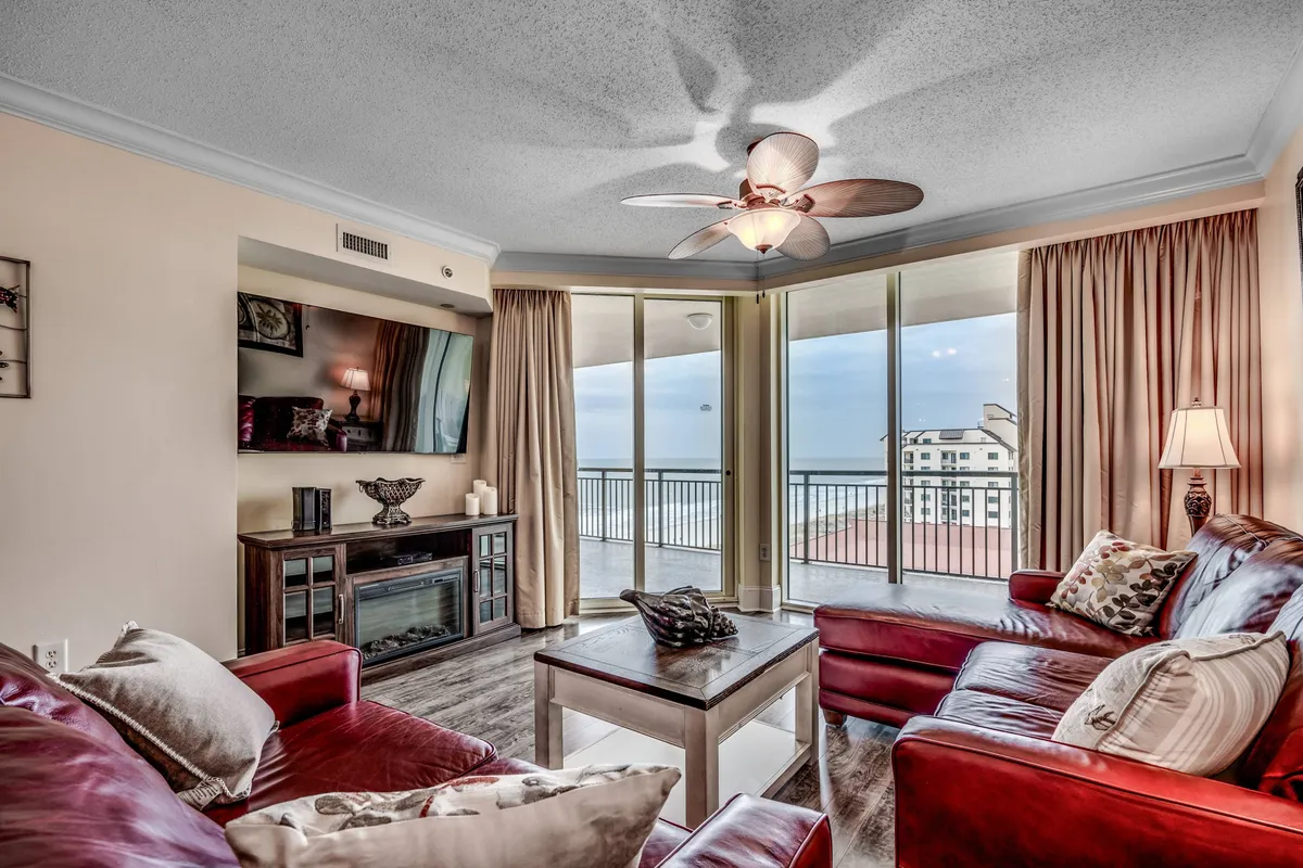 A cozy living room featuring rich red leather sofas, a stylish wooden coffee table, a ceiling fan, and large windows that showcase sweeping ocean vistas. The space feels perfect for relaxing, watching TV, or hosting a game night with friends and family.