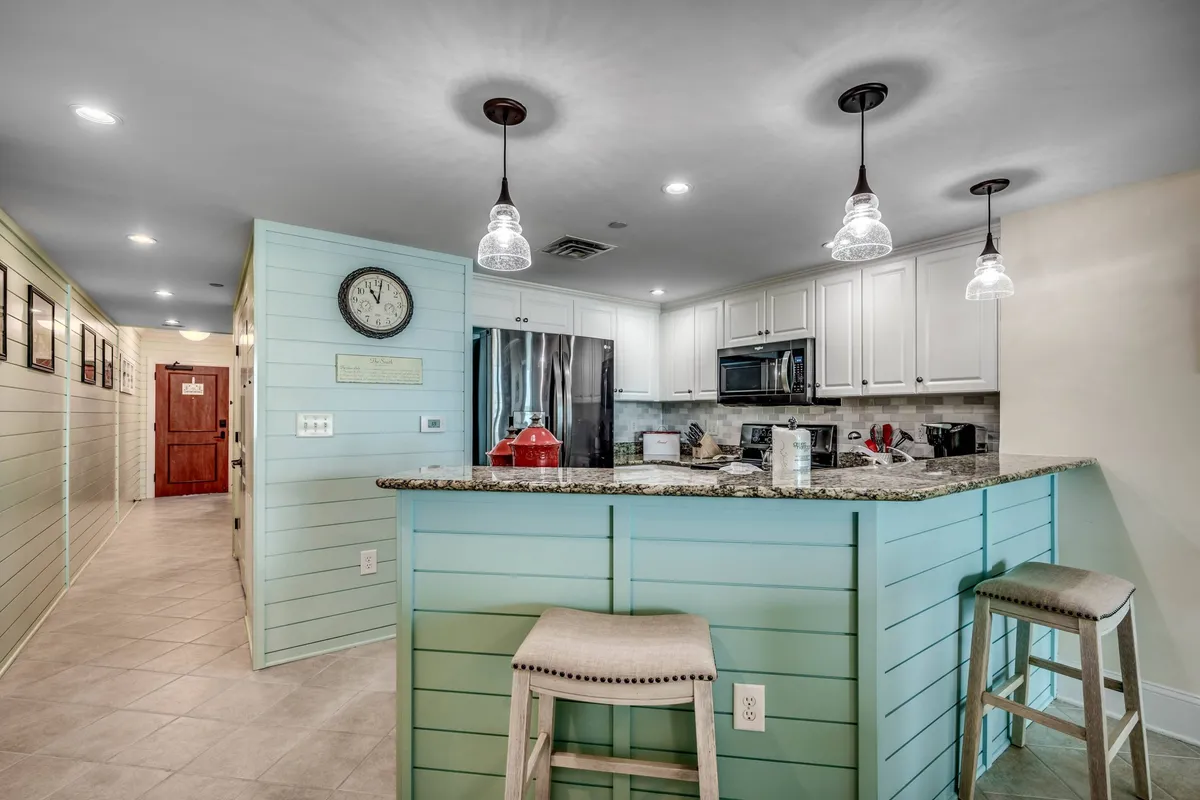 A bright, inviting kitchen with a spacious granite island, stylish pendant lighting, and a large wall clock. The space is arranged for group gatherings, perfect for cooking, socializing, or enjoying a relaxed morning coffee.