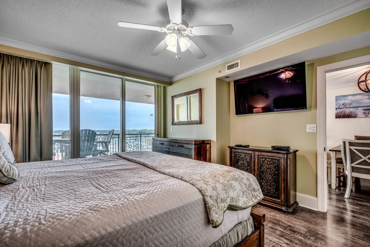 A serene bedroom with a neatly made bed facing a private balcony, offering an inviting view of the ocean beyond. The room is decorated with tasteful furniture and includes a television, blending relaxation with modern comforts.