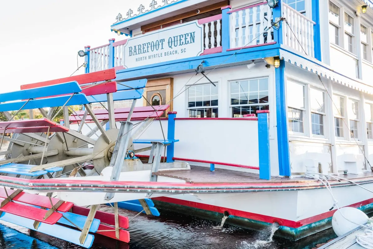 A vibrant exterior shot of a paddle wheel boat with a colorful facade and prominent signage, capturing the festive mood of a riverboat dinner cruise experience. The image hints at sunset gatherings, social dining, and lively entertainment on the water.