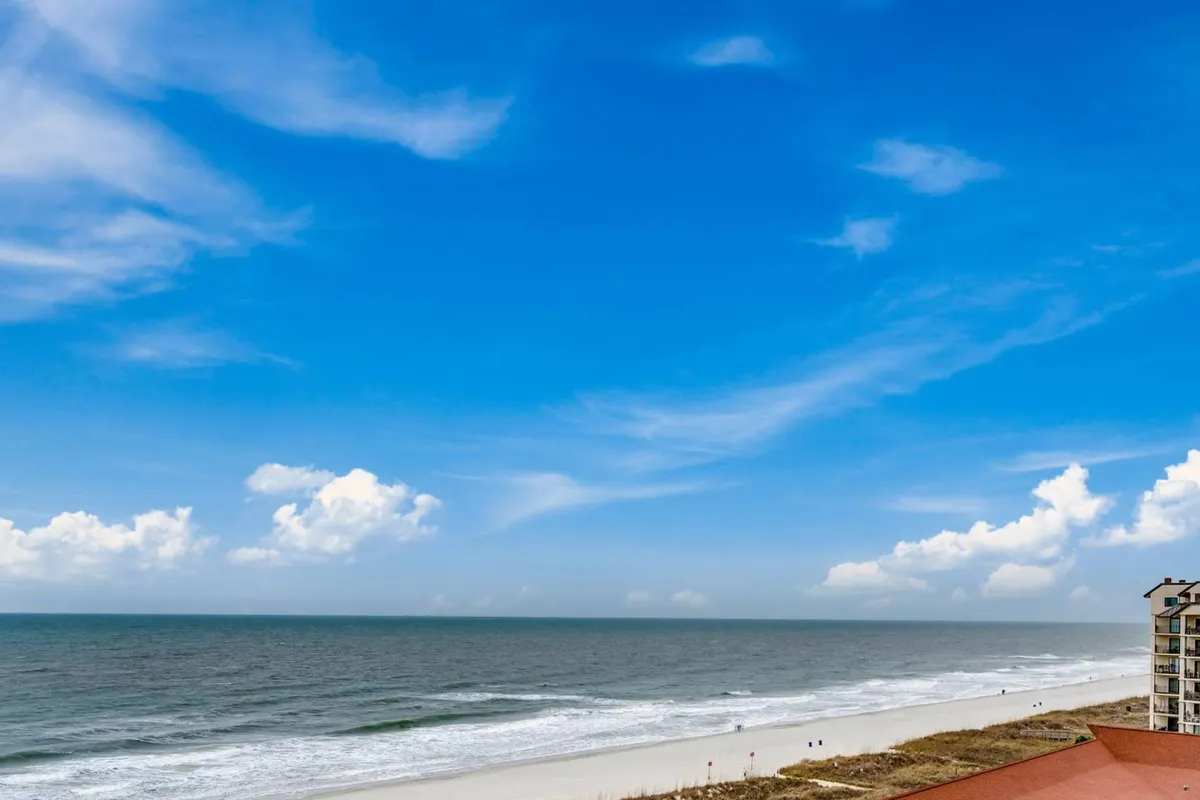 A stunning beachfront view with a wide expanse of sandy shoreline, gentle ocean waves, and a few people enjoying activities like walking, sunbathing, and swimming. The sky is bright with clouds, and the Mar Vista Grande building is visible, situating the viewer amidst the lively, family-friendly atmosphere of North Myrtle Beach.