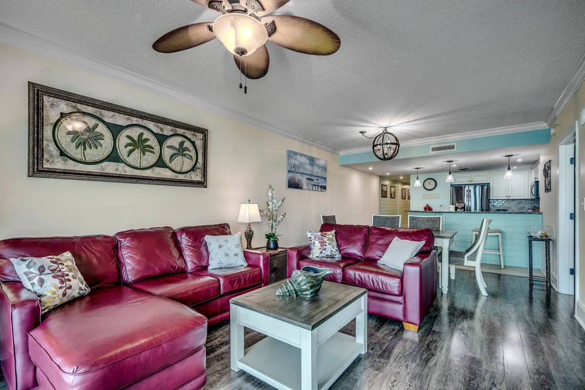 This image shows a cozy interior living area with red leather sofas, decorative wall art, a coffee table with decorative items, and a kitchen area visible in the background. The ambiance suggests a space for relaxation, socializing, and enjoying home-like comforts within a resort setting.