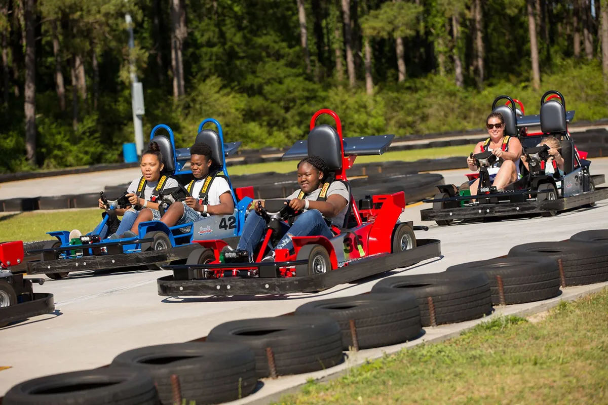 A lively exterior shot of a go-kart track, with several people enjoying a friendly and competitive race. The scene is filled with energy and excitement, featuring participants, track barriers, and the sense of dynamic family activity.