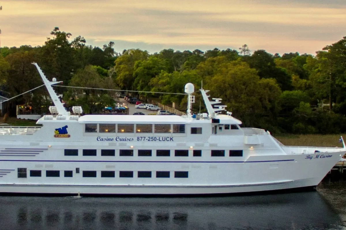 A large, elegant white casino cruise boat glides along the water, surrounded by lush greenery and a distant shoreline. People are seen enjoying dining, entertainment, and scenic views from the deck, basking in the vibrant yet relaxed atmosphere.