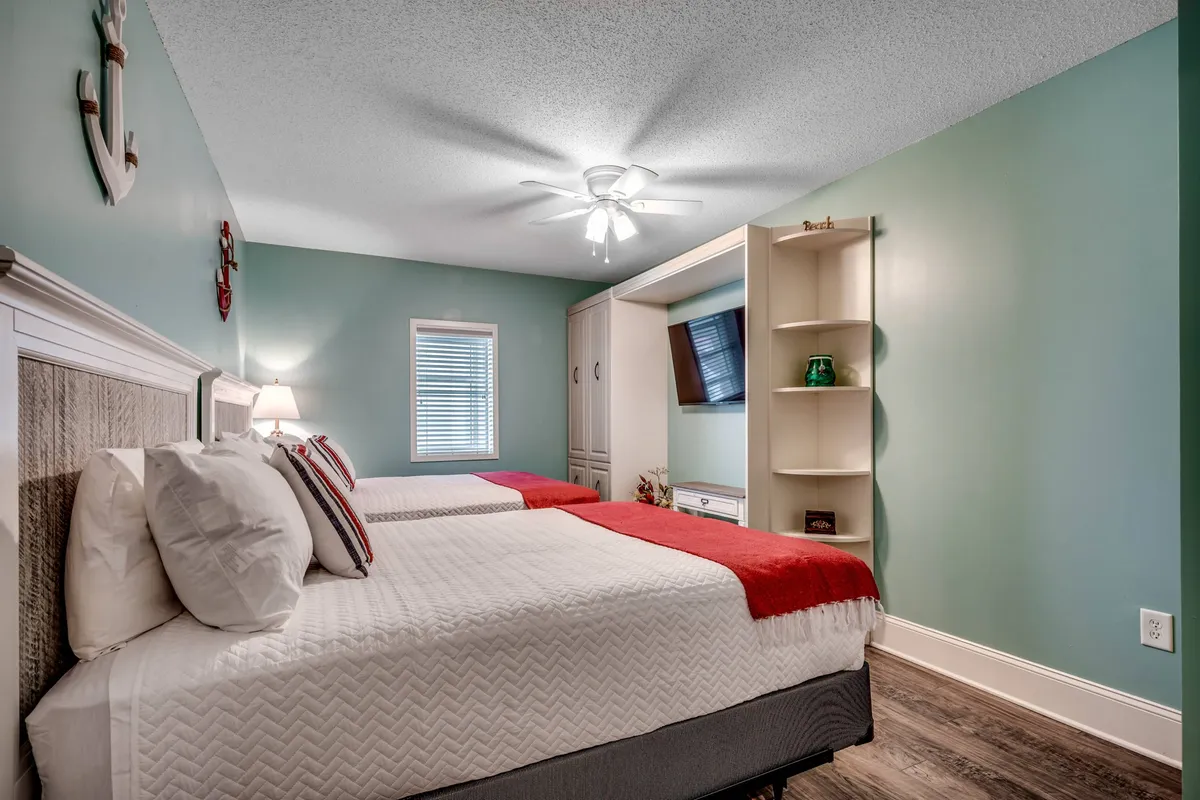 The image shows a guest bedroom with two comfortable beds featuring red accents, built-in shelving, and a window letting in ample natural light. The space radiates comfort and provides a welcoming environment for guests to relax, read, or enjoy TV.