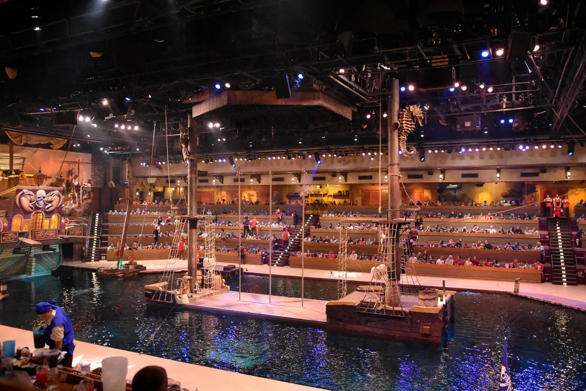 This image showcases a vibrant interior space with a large pirate ship structure, audience seating, and a water area with boats. The scene is lively, suggesting entertainment and community events available within the resort.